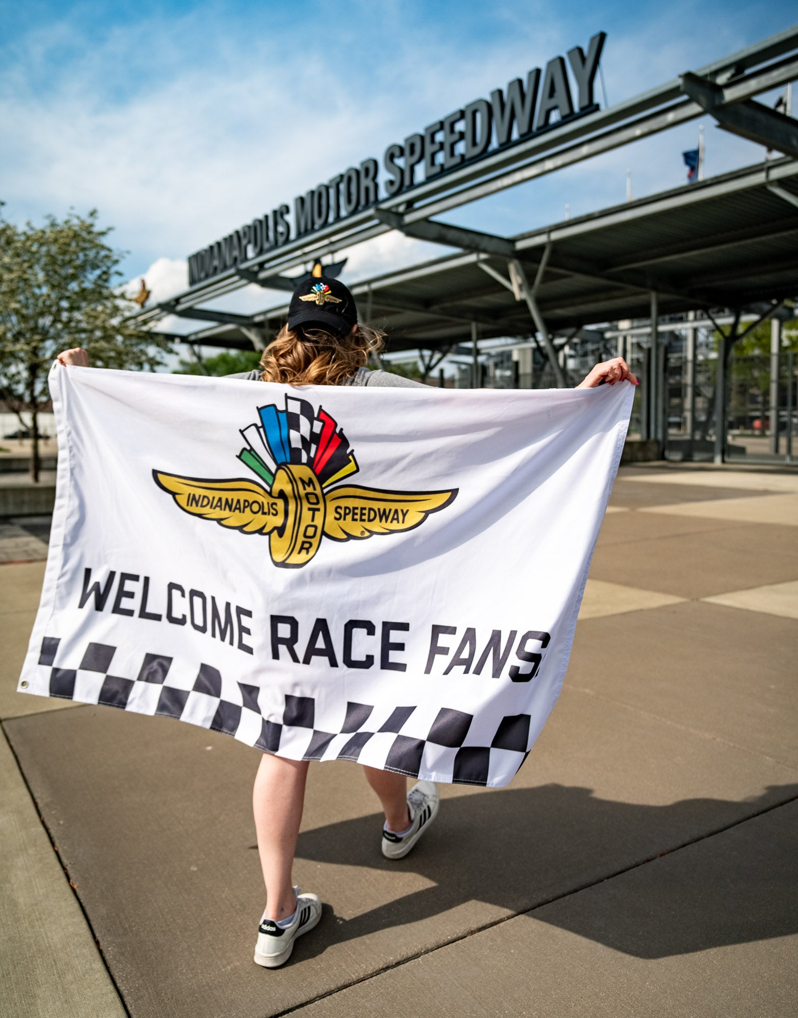 An Indianapolis Motor Speedway "Welcome Race Fans" flag being held by a woman walking into the IMS main gate.