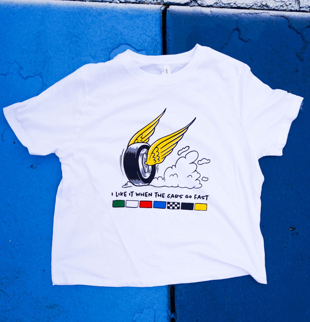 White t-shirt with wing and wheel racing graphic and text "I like it when the cars go fast" on a blue background