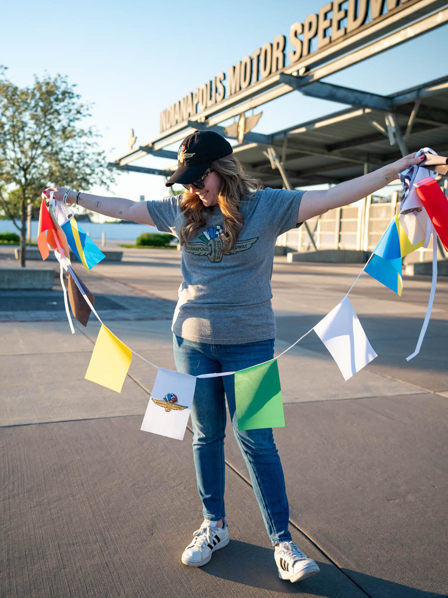 An Indianapolis Motor Speedway "Welcome Race Fans" racing flag bunting being held by a woman in front of the IMS main gate.