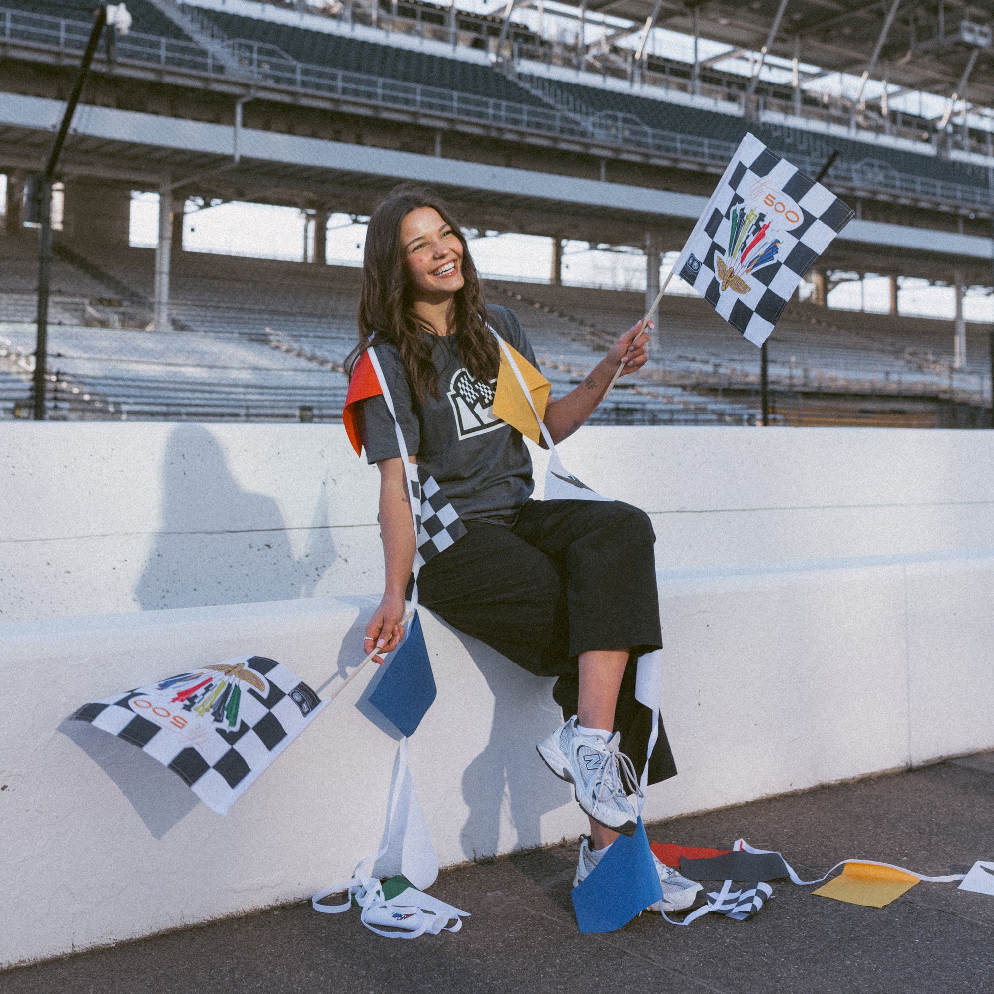 woman at race track draped with racing flag bunting while smiling and holding limited edition vintage logo checkered flags in both hands 