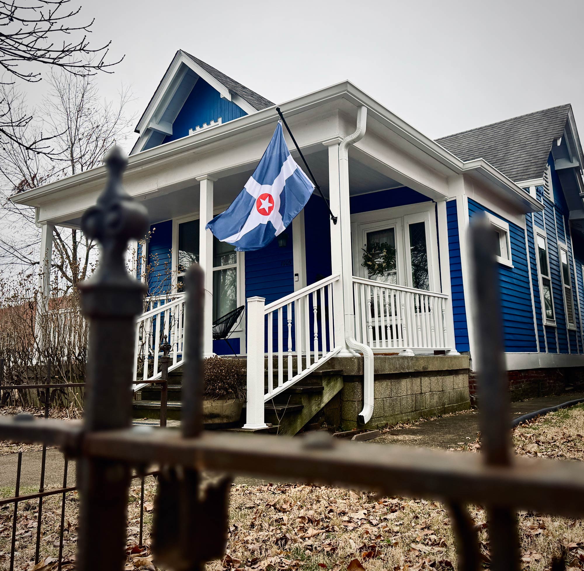 Blue house with a white porch and an Indianapolis flag, seen through a metal fence.