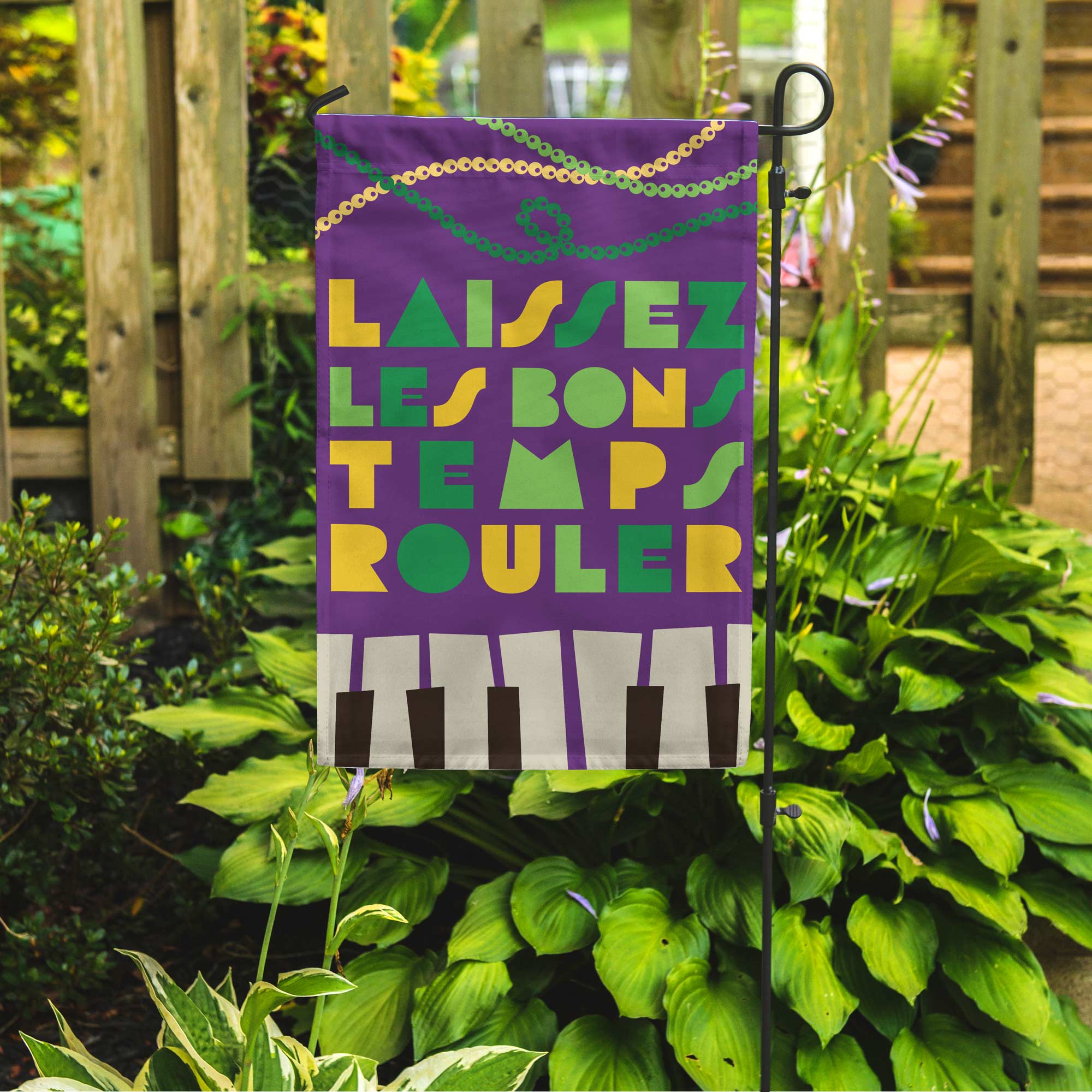 Decorative garden flag with colorful text reading "Laiessez Les Bons Temps Rouler" and a piano design, attached to a stand among plants.