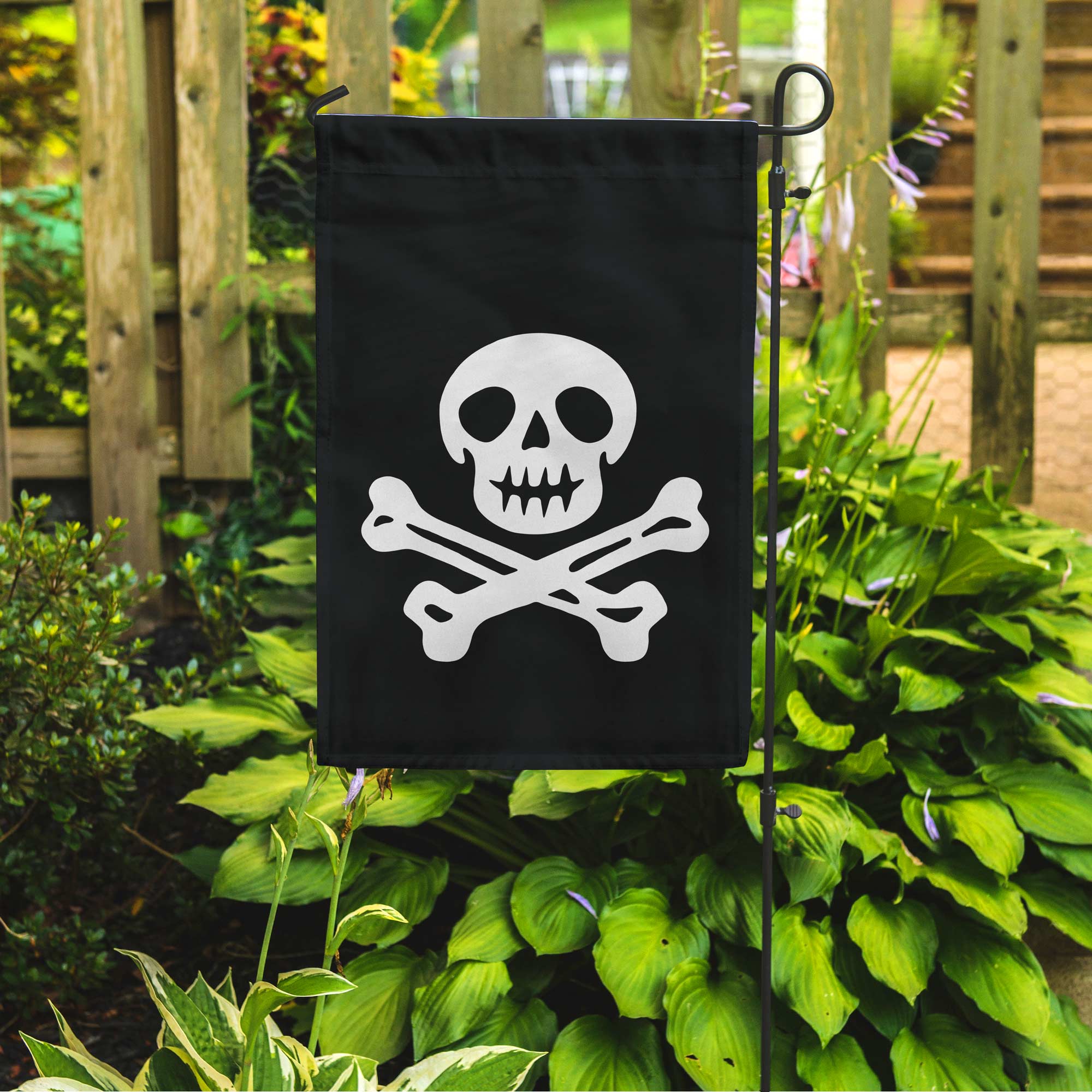 Black garden flag with a white skull and crossbones design, attached to a stand in a garden setting.
