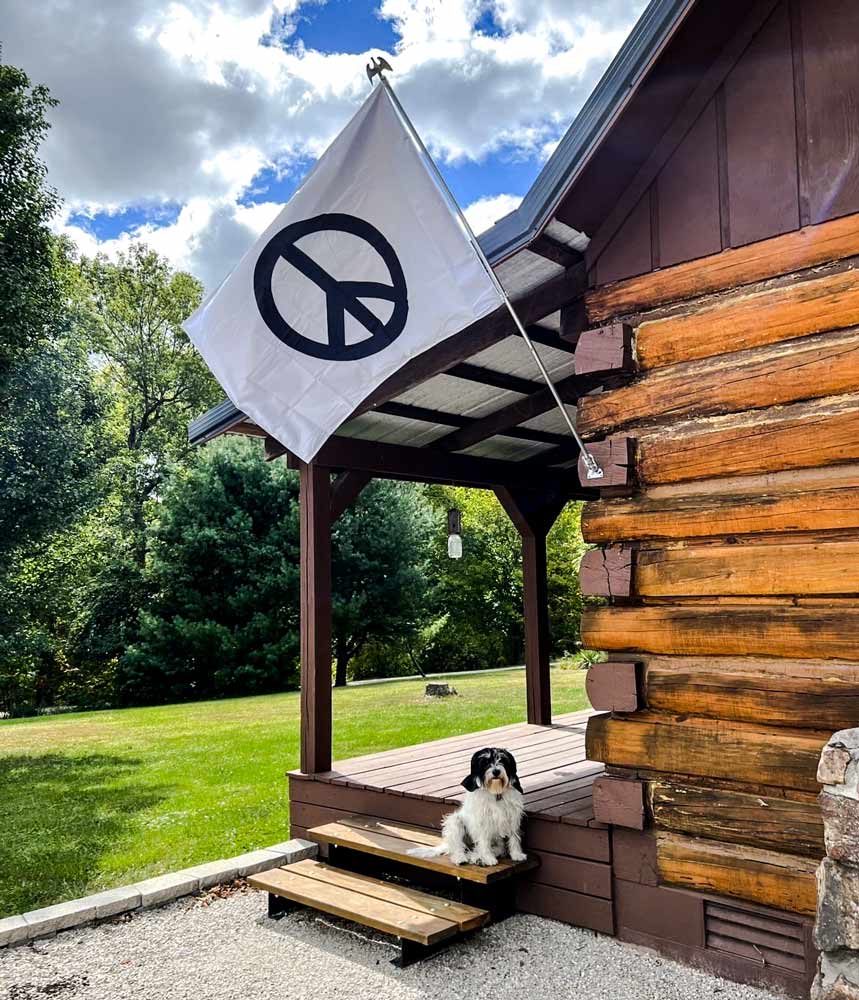 White peace sign flag on the side of a log cabin with a dog on the porch
